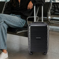 Black cabin trolley suitcase with four wheels, near seated person in terminal.